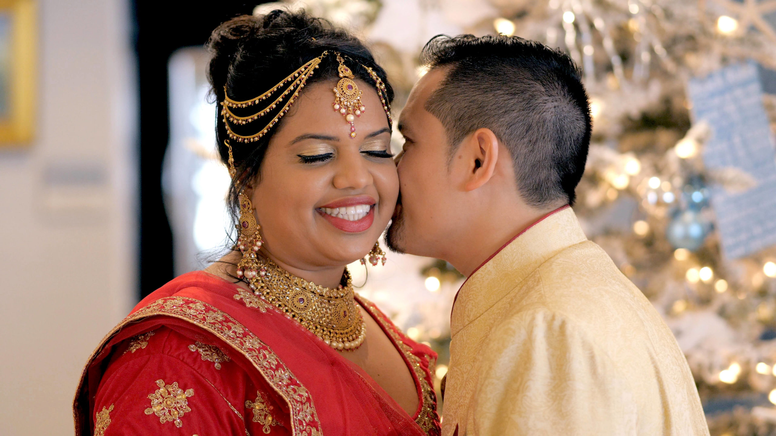Filipino groom whispering in Indian bride's ear in front of a Christmas tree