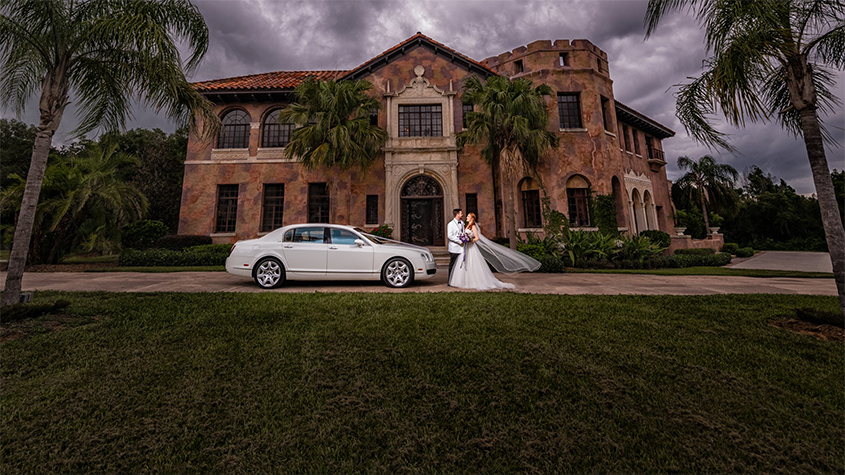 Married couple standing next to a luxury car in front of The Howey Mansion