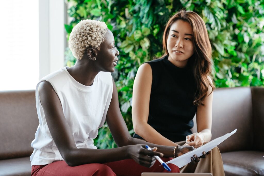 Black woman holding a pen and paper having a conversation with an Asian women as they sit on a couch
